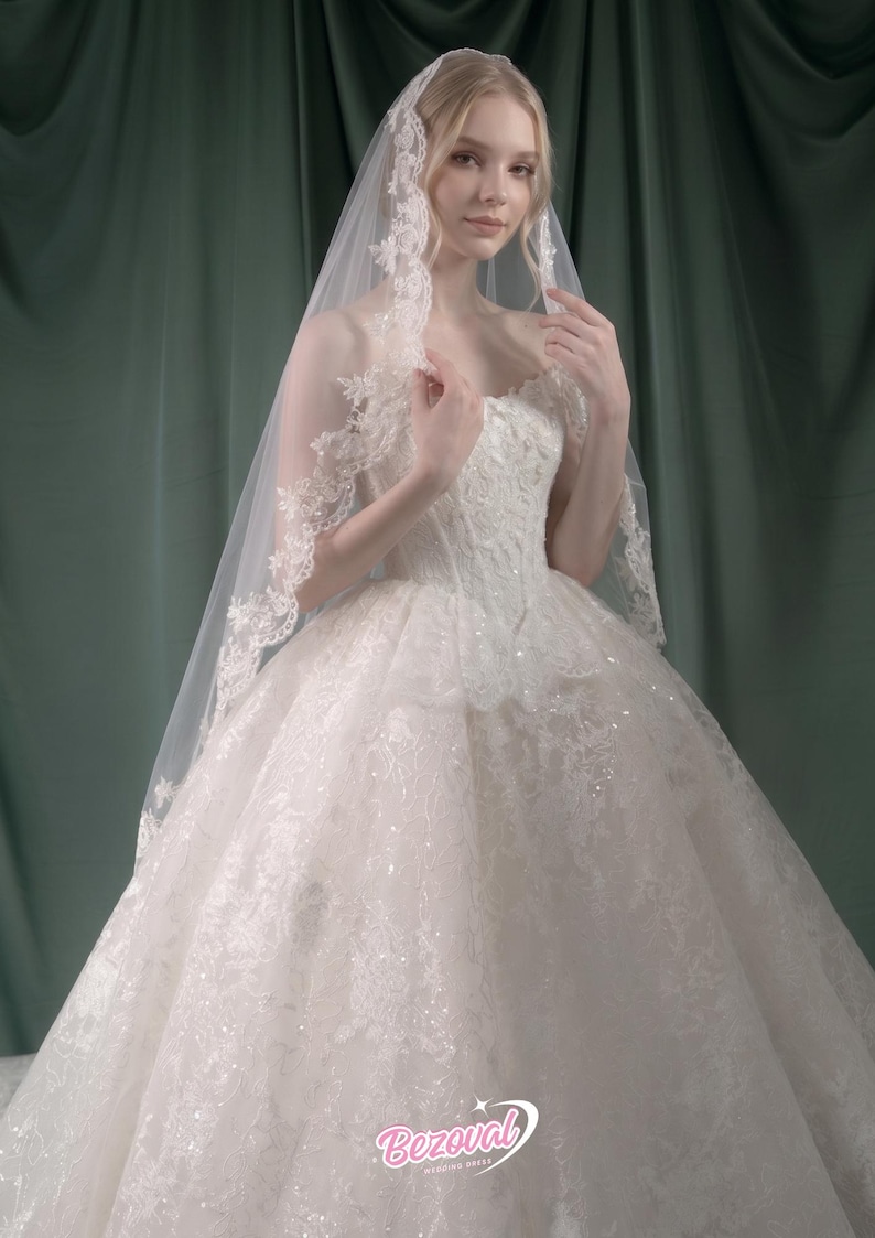 Puede incluir: Vestido de novia blanco con falda amplia y corpi&ntilde;o detallado. El vestido est&aacute; adornado con encaje y lentejuelas. Un velo transparente con ribete de encaje cae sobre los hombros. Logo "Bezoval Wedding Dress".