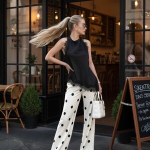 May include: A black halter top with lace trim is paired with white palazzo pants featuring large black polka dots. A white handbag and sunglasses complete the outfit. The image is taken outside a cafe.