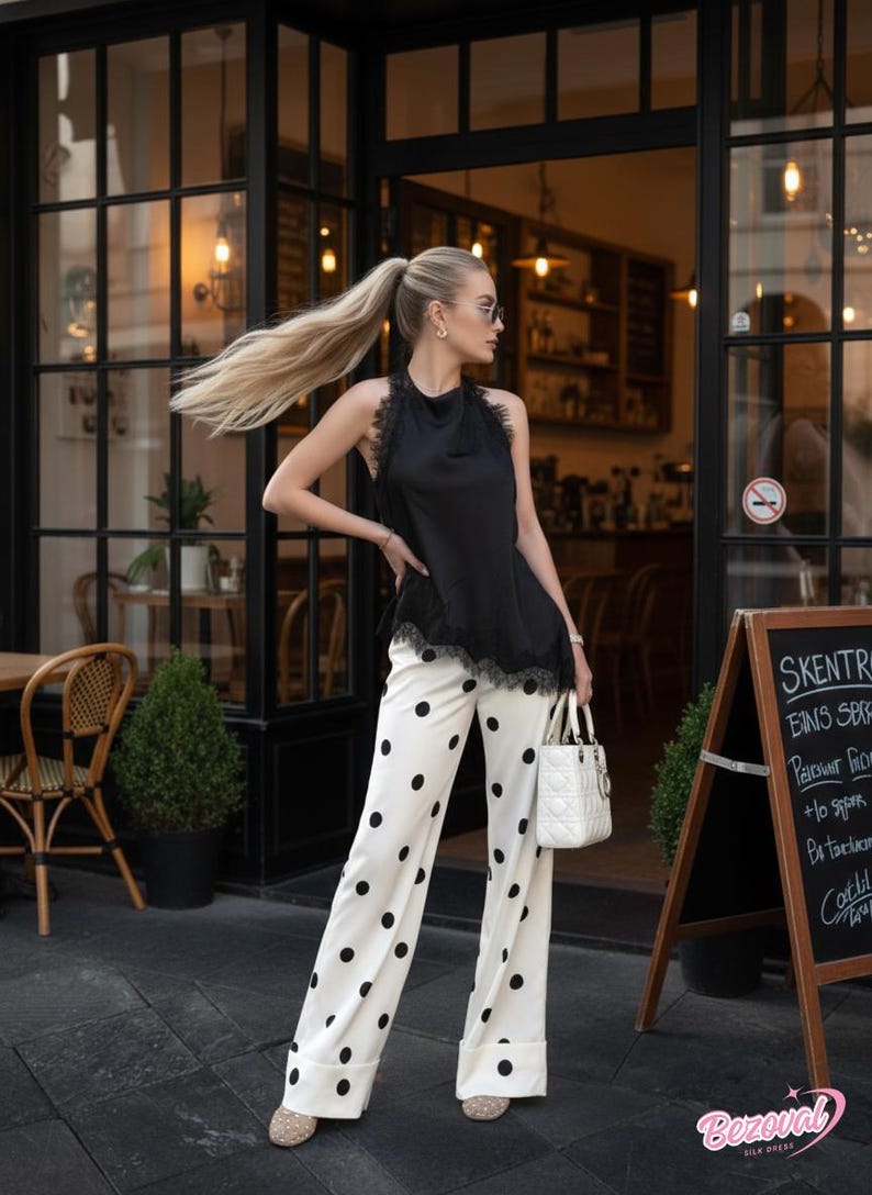 May include: A black halter top with lace trim is paired with white palazzo pants featuring large black polka dots. A white handbag and sunglasses complete the outfit. The image is taken outside a cafe.