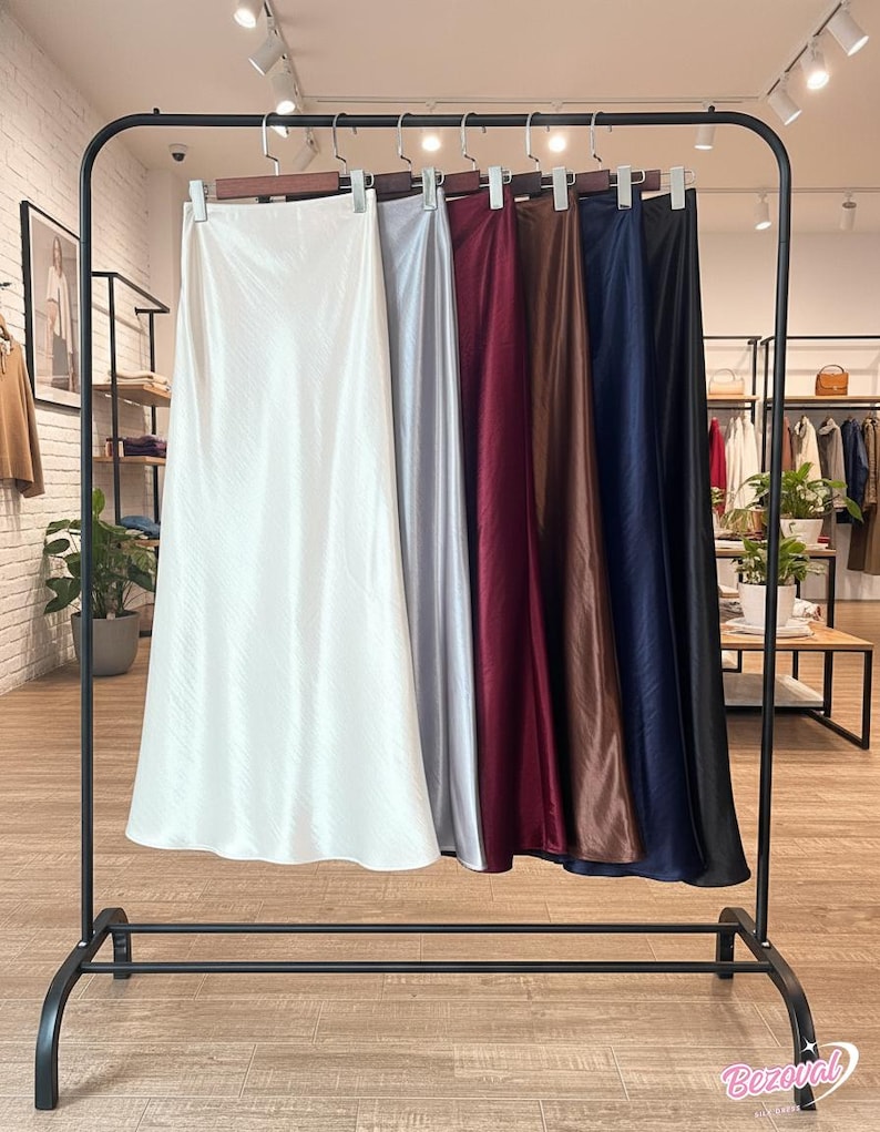 May include: A clothing rack displays several satin midi skirts in various colors, including white, silver, burgundy, brown, navy, and black. The skirts hang from wooden hangers on a black metal rack, showcasing their smooth, flowing fabric.