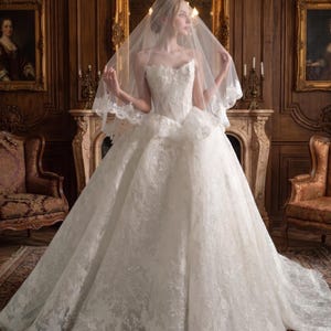 Puede incluir: Un vestido de novia blanco, sin tirantes, con una falda amplia y detalles de encaje intrincados. La novia lleva un velo con ribete de encaje. El vestido es de Bezoval Wedding Dresses.