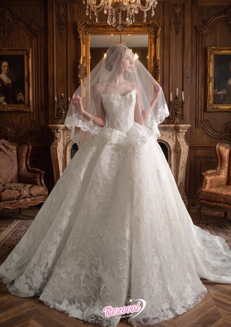 Puede incluir: Un vestido de novia blanco, sin tirantes, con una falda amplia y detalles de encaje intrincados. La novia lleva un velo con ribete de encaje. El vestido es de Bezoval Wedding Dresses.