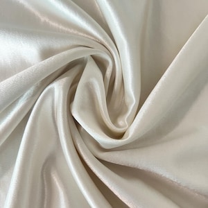May include: Close-up of a cream-colored satin fabric with a lustrous sheen. The fabric is draped in soft folds, creating shadows and highlights. The texture appears smooth and flowing, suggesting a luxurious material.