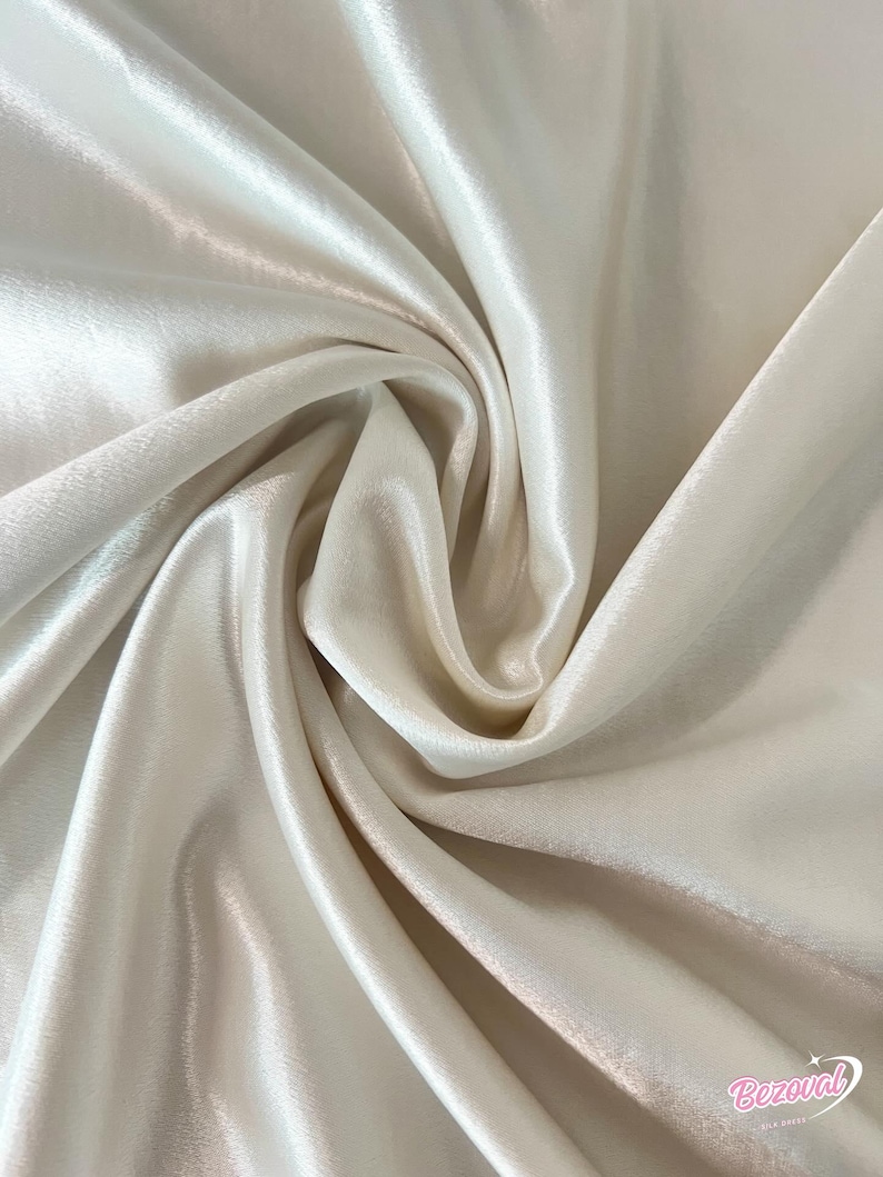 May include: Close-up of a cream-colored satin fabric with a lustrous sheen. The fabric is draped in soft folds, creating shadows and highlights. The texture appears smooth and flowing, suggesting a luxurious material.