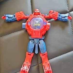 May include: A red and blue plastic toy robot with a Spider-Man design. The robot has a large head with a Spider-Man mask and a clear plastic window on the chest with the words "Spider-Man" printed on it.