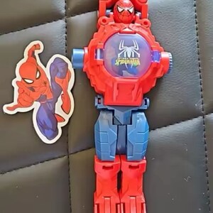 May include: A red and blue plastic toy watch with a Spider-Man design. The watch face has a clear plastic cover with a Spider-Man image inside. The watch has a red strap with a blue buckle. The watch is accompanied by a sticker of Spider-Man.
