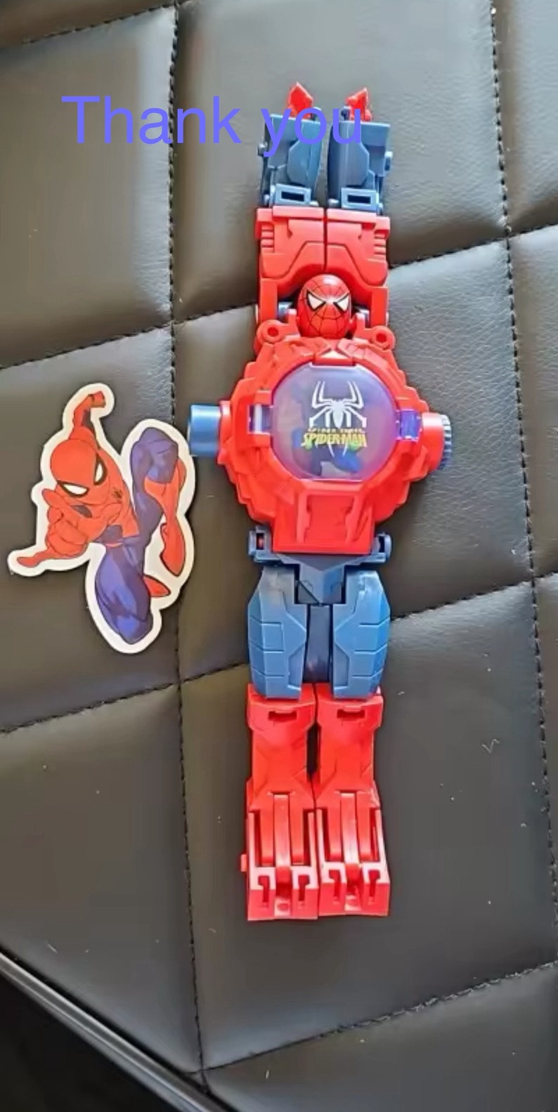 May include: A red and blue plastic toy watch with a Spider-Man design. The watch face has a clear plastic cover with a Spider-Man image inside. The watch has a red strap with a blue buckle. The watch is accompanied by a sticker of Spider-Man.