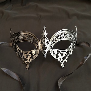 May include: A silver-colored, filigree masquerade mask with intricate cut-out designs. The mask has a central decorative element and is attached to black ribbons. The mask is designed to cover the eyes and is suitable for a masquerade ball or costume party.