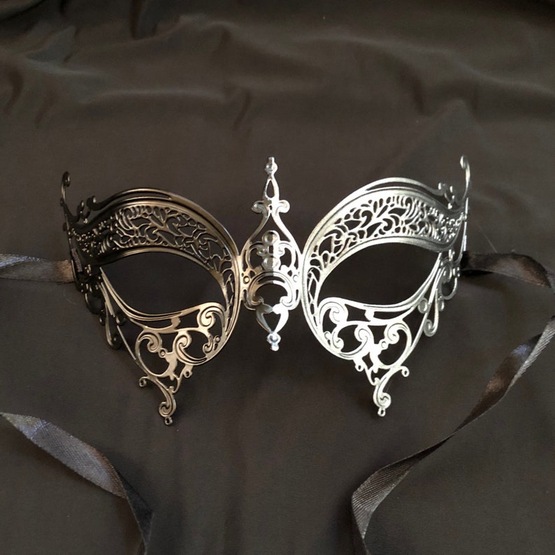 May include: A silver-colored, filigree masquerade mask with intricate cut-out designs. The mask has a central decorative element and is attached to black ribbons. The mask is designed to cover the eyes and is suitable for a masquerade ball or costume party.