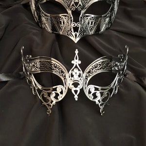 May include: Two silver masquerade masks with intricate, delicate designs. The masks are made of metal and have a black background.