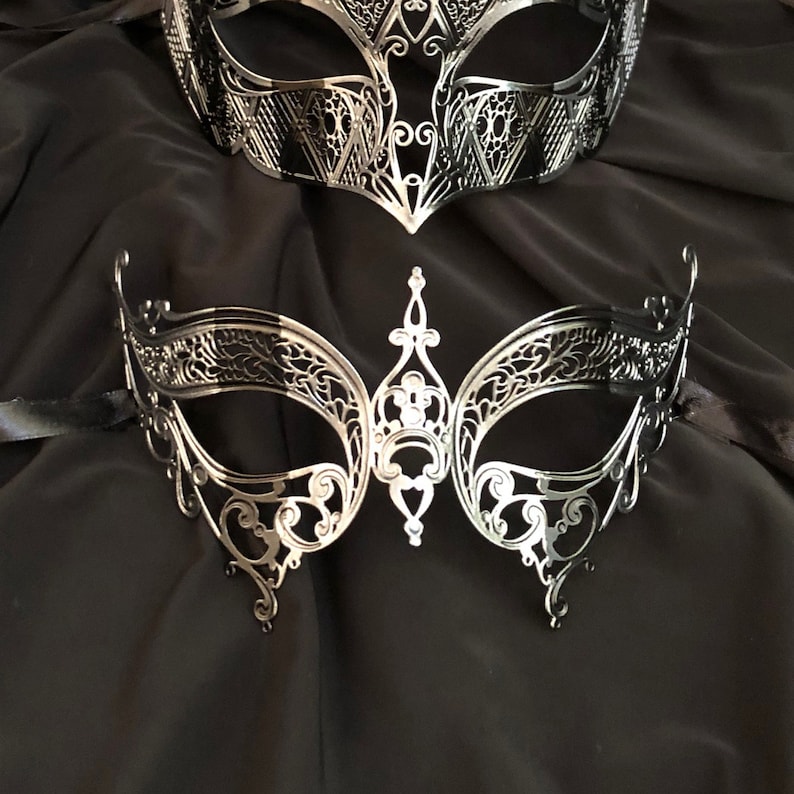 May include: Two silver masquerade masks with intricate, delicate designs. The masks are made of metal and have a black background.