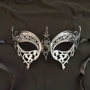 May include: Silver-colored masquerade mask with intricate filigree detailing. The mask features a central decorative element and is attached to black satin ribbons. The mask is designed to cover the eyes and upper face.