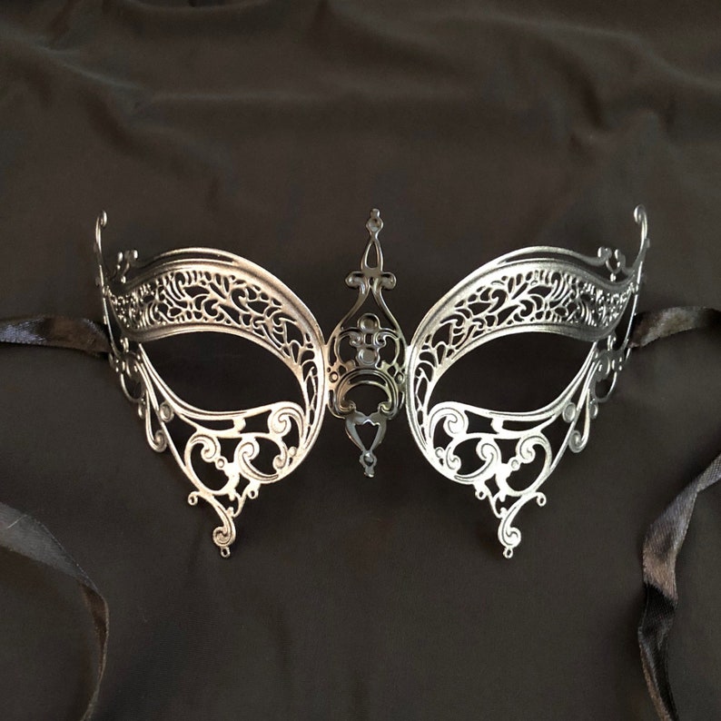 May include: Silver-colored masquerade mask with intricate filigree detailing. The mask features a central decorative element and is attached to black satin ribbons. The mask is designed to cover the eyes and upper face.