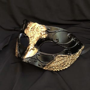 Könnte beinhalten: Eine schwarz-goldene Maske für einen Maskenball mit aufwendigen Details. Die Maske hat eine schwarze Basis mit goldenen Akzenten, darunter verzierte Muster und ein strukturiertes Flügeldesign. Die Augenöffnungen sind oval, und die Maske hat ein schwarzes Band zum Befestigen.