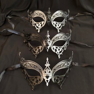 May include: Three ornate, filigree-style masquerade masks in silver and black. Each mask features a symmetrical design with intricate cut-out patterns and attached black ribbons. The masks are displayed on a dark, draped fabric background.