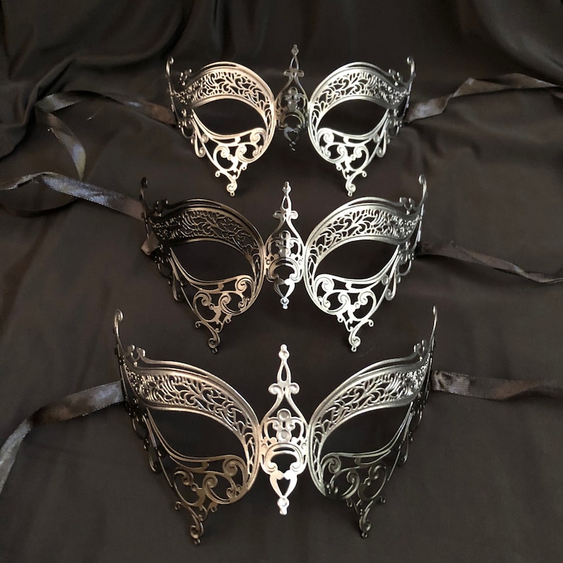 May include: Three ornate, filigree-style masquerade masks in silver and black. Each mask features a symmetrical design with intricate cut-out patterns and attached black ribbons. The masks are displayed on a dark, draped fabric background.