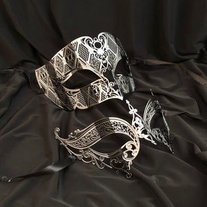 May include: Two silver masquerade masks with intricate designs. The masks are on a black fabric background.