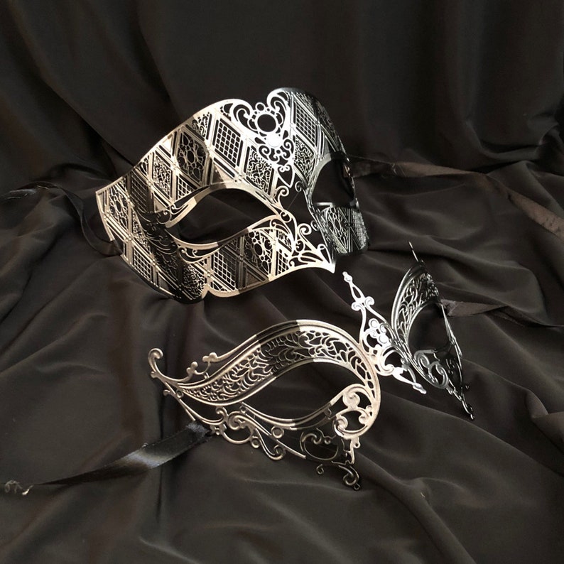 May include: Two silver masquerade masks with intricate designs. The masks are on a black fabric background.
