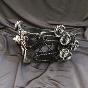 May include: A black steampunk-style mask with intricate gear details and a black leather strap. The mask has a lace-like design and is decorated with silver gears.