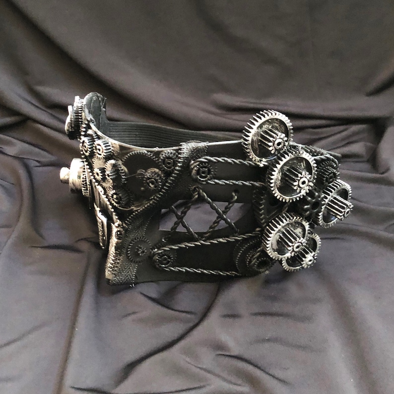 May include: A black steampunk-style mask with intricate gear details and a black leather strap. The mask has a lace-like design and is decorated with silver gears.