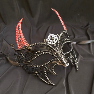 May include: A black masquerade mask with red horns and black rhinestones. The mask has a delicate, intricate design and is perfect for a masquerade ball or costume party.