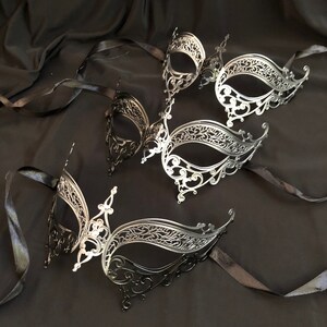 May include: An assortment of ornate, filigree-style masquerade masks in silver and black. Each mask features intricate cut-out designs and is attached to a black satin ribbon. The masks are arranged on a dark, textured background.