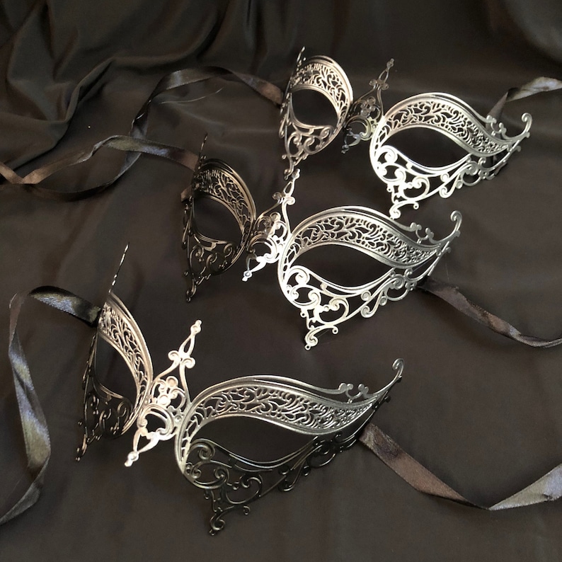 May include: An assortment of ornate, filigree-style masquerade masks in silver and black. Each mask features intricate cut-out designs and is attached to a black satin ribbon. The masks are arranged on a dark, textured background.