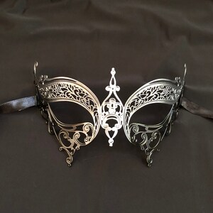 May include: A silver-colored masquerade mask with intricate filigree detailing. The mask features a central decorative element and is attached to a black ribbon. The design is symmetrical and elegant, suitable for formal events.