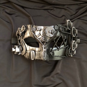 May include: A silver and black masquerade mask with a steampunk design featuring gears and cogs. The mask has a black leather strap.