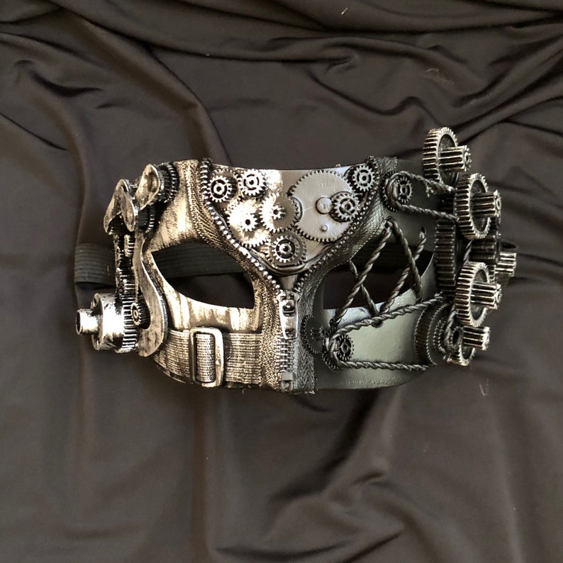 May include: A silver and black masquerade mask with a steampunk design featuring gears and cogs. The mask has a black leather strap.