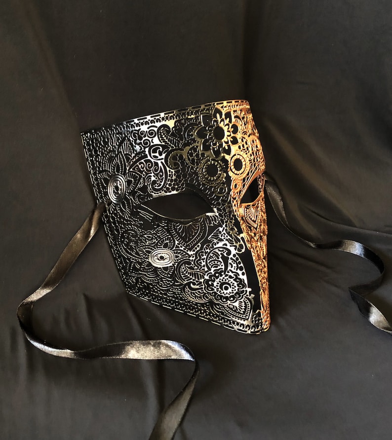 May include: A black and gold masquerade mask with intricate filigree designs. The mask has a single eye hole and is attached to black satin ribbons.