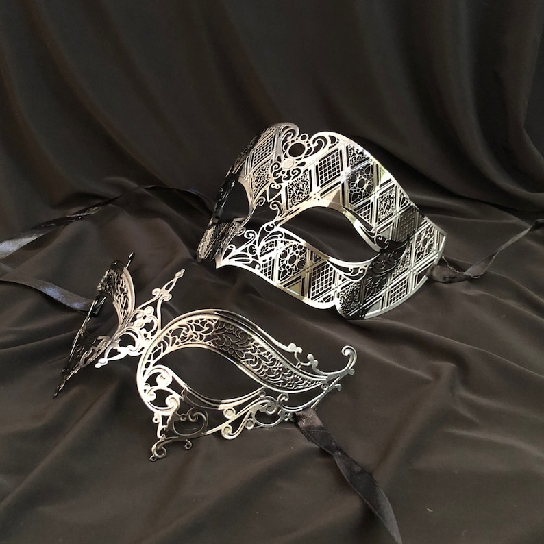 May include: Two silver masquerade masks with intricate designs. One mask is a traditional eye mask with a diamond pattern, the other is a more elaborate design with a flowing shape.