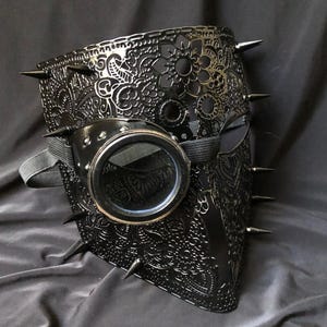 May include: Black steampunk mask with floral filigree detailing, metal spikes, and a single round goggle. The mask features an elastic strap. The mask is made of metal.