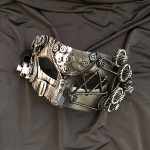 May include: A silver and black masquerade mask with a steampunk design featuring gears and cogs. The mask has a black strap and is made of plastic.