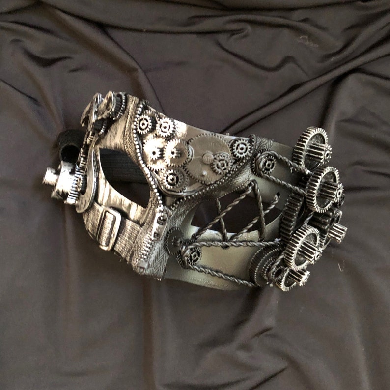 May include: A silver and black masquerade mask with a steampunk design featuring gears and cogs. The mask has a black strap and is made of plastic.
