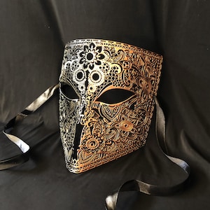 May include: A gold and black masquerade mask with intricate floral designs. The mask has a black ribbon attached to the sides.