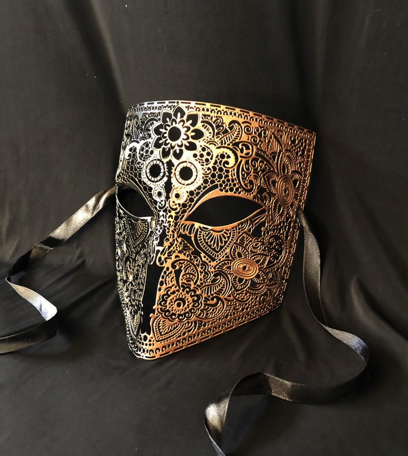 May include: A gold and black masquerade mask with intricate floral designs. The mask has a black ribbon attached to the sides.