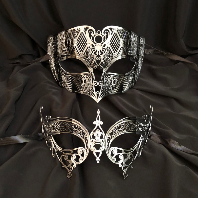 May include: Two silver masquerade masks with intricate designs. The top mask has a diamond pattern and the bottom mask has a more floral design.