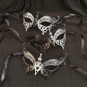 May include: An assortment of ornate masquerade masks in silver and black, arranged on a dark, textured fabric. The masks feature intricate filigree designs and are tied with black ribbons, suggesting a theme of elegance and mystery.