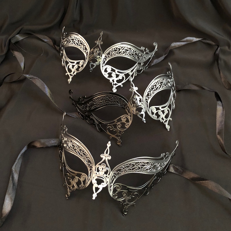 May include: An assortment of ornate masquerade masks in silver and black, arranged on a dark, textured fabric. The masks feature intricate filigree designs and are tied with black ribbons, suggesting a theme of elegance and mystery.