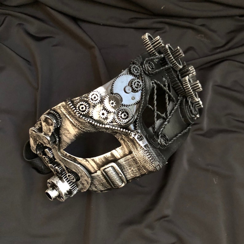 May include: A silver and black masquerade mask with a steampunk design. The mask features gears, cogs, and a leather strap.