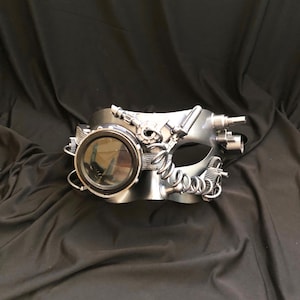 May include: A silver steampunk-style masquerade mask with a round, clear lens and intricate gears and pipes.