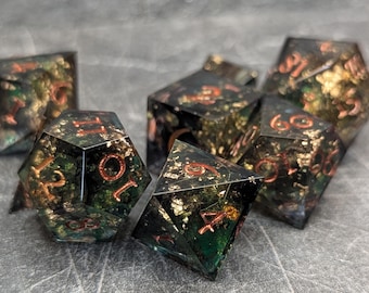 May include: A set of polyhedral dice with a dark, speckled appearance. The dice have a mix of green, gold, and copper tones, with copper-colored numbers. The dice are designed for tabletop gaming.