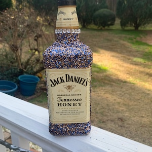 May include: A decorated Jack Daniel's Tennessee Honey bottle. The bottle is covered in blue and gold glitter, with a band of clear rhinestones near the top. The label reads "Jack Daniel's Tennessee Honey".