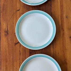 May include: Three white ceramic plates with a light blue border are arranged vertically on a wooden surface. The plates are round and have a simple, clean design, suitable for everyday use.