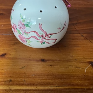 May include: A white ceramic sphere decorated with pink roses, green leaves, and pink ribbons. It features three small holes and a pink cord. The sphere is a decorative item.