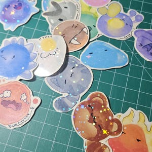 May include: A collection of assorted holographic stickers featuring various cartoon creatures, including a brown bear, a blue sun, a pink blob, and a gray cat. Each sticker has a glossy finish with a star-shaped holographic effect that reflects light.