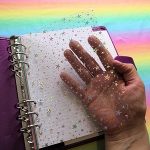 Może przedstawiać: Przezroczysta folia plastikowa z holograficznym wzorem gwiazd umieszczona jest na fioletowym segregatorze z srebrnym mechanizmem pierścieniowym. Segregator ma napis "FILOFAX" nadrukowany na grzbiecie.