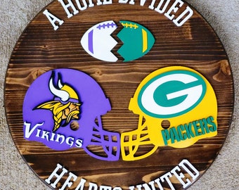 May include: A wooden sign with the text "A Home Divided Hearts United" in white letters. The sign features a purple and yellow football helmet with a Viking logo and a green and yellow football helmet with a Packers logo. The sign also features a broken football with purple and green colors.