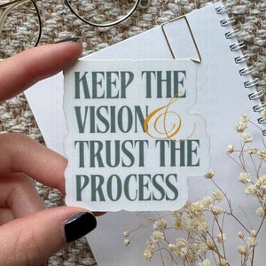 May include: A white sticker with the text "KEEP THE VISION TRUST THE PROCESS" in dark green, with a gold flourish. The sticker is held by a hand with black nail polish, near a notebook, glasses, and dried flowers.